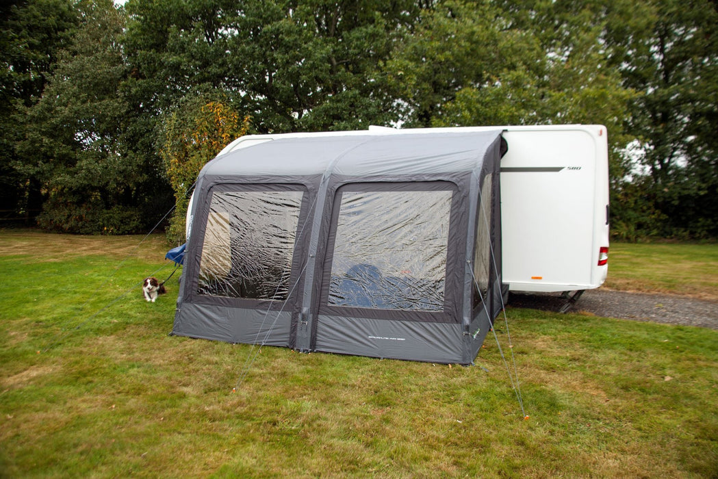 Outdoor Revolution Sport Lite Air 330 Inflatable Caravan Awning - main product image, showing front-right side view of awning attached to caravan pitched in field with surrounding trees, and dog lying on grass to left-hand side of awning