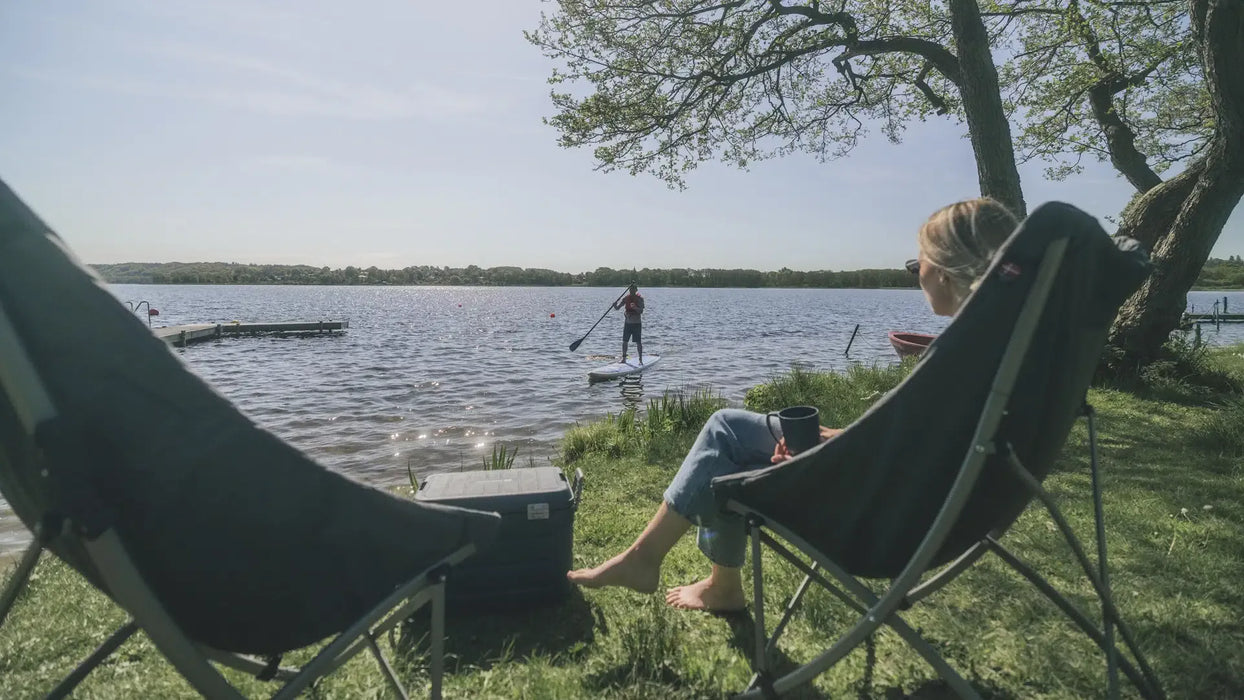 Outwell Tally Lake Camping Chair - lifestyle image, showing person sat in chair with a second empty chair to their left, a coolbox in between the two, and a person paddleboarding on water in the near distance