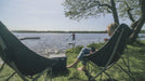 Outwell Tally Lake Camping Chair - lifestyle image, showing person sat in chair with a second empty chair to their left, a coolbox in between the two, and a person paddleboarding on water in the near distance