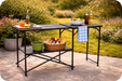 Quest Rosemary Folding Camping Kitchen Stand – lifestyle image, showing unit in use with food and towel on it, against garden background with patio