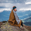 Robens Icefall Quilt - Camping Blanket main feature image lifestyle image of lady with blanket over her sat on rock with snowing mountains in background