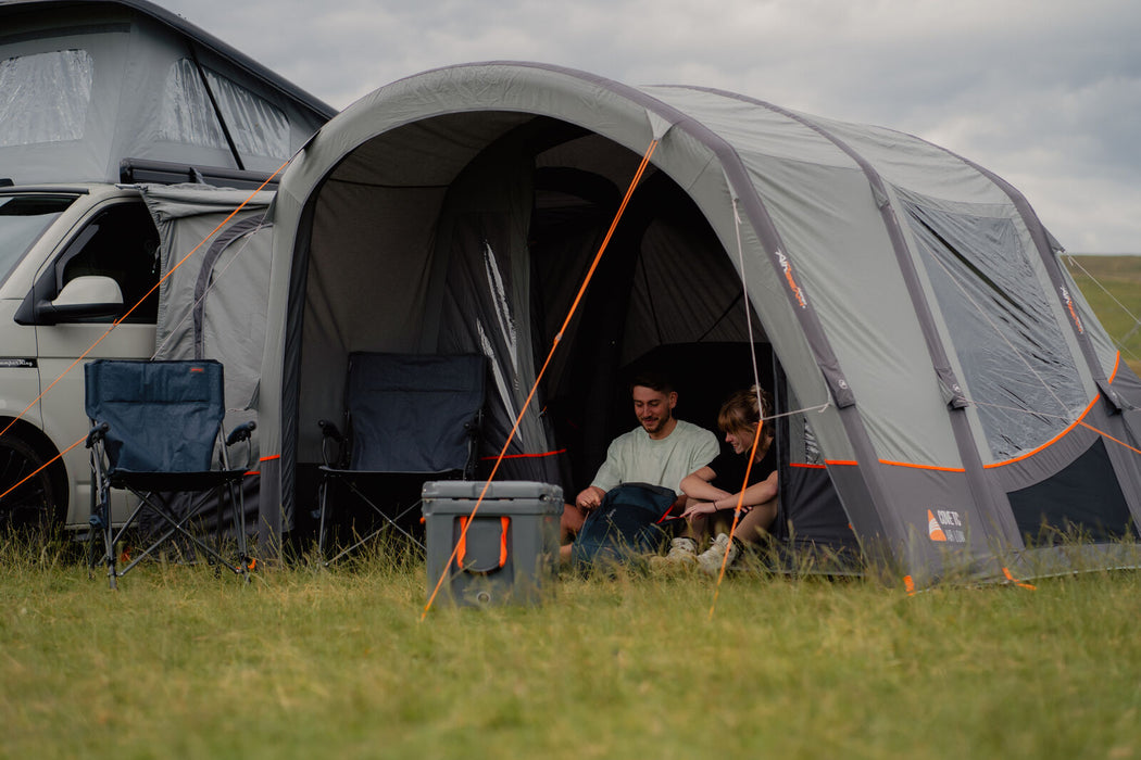 Vango Cove Air TC Inflatable Drive Away Awning Grey - Low lifestyle image of awing pitched against vw pop top with close up of awning canopy front with door open. couple sitting in the front entrance with coolbox and chairs out front.