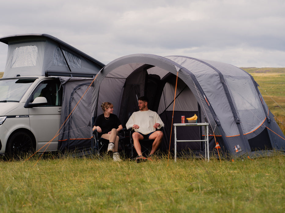 Vango Cove III Air Inflatable Drive Away Awning Smoke Grey - Low front lifestyle view of awning attached to pop top van on left side, a couple sat in canopy with table to the right with bananas on. in field with hill behind