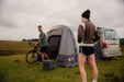 Vango Faros III Air Inflatable Drive Away Awning Cloud Grey - Low side view from rear of van with woman in foregrounds and man leabing awning iwth bike. coolbox on the grass and privacy curtains toggled up. in field with hill in background