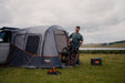 Vango Faros III Air Inflatable Drive Away Awning Cloud Grey - Low lifestyle image of awning showing left side entrances with front entrance unzipped. privacy curtains over side door up. man exiting entrance with a bike. shoes and coolbox on ground outside. lake in background 