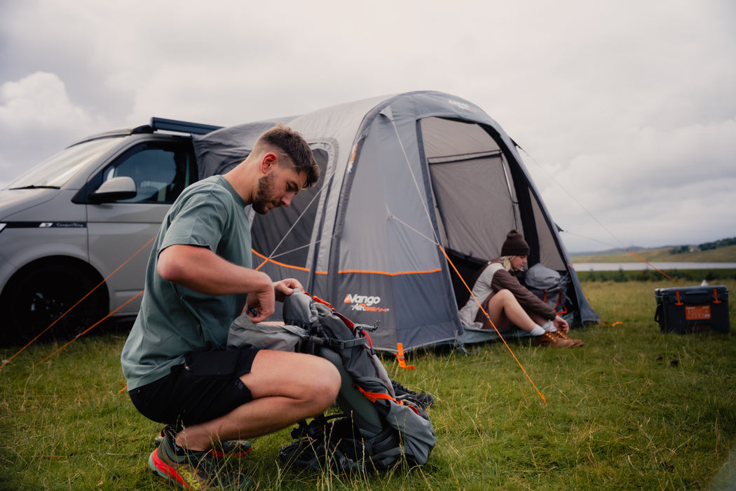 Vango Faros III Air Inflatable Drive Away Awning Cloud Grey - Low lifestyle image with man in foreground and woman in awning entrance, lake behind and awning attached to van 