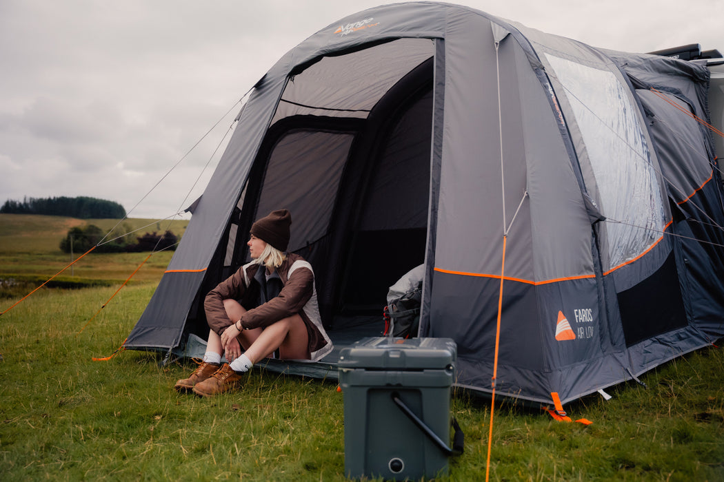 Vango Faros III Air Inflatable Drive Away Awning Cloud Grey - Low front  angled view of awning with van behind. back door zipped up and a woman sat in front entrance with hill in background. coolbox on ground in front