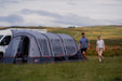 Vango Galli CC Air Inflatable Drive Away Awning - mid lifestyle image of awning side view attached to van with front door half open. grass field in front and hills in background. women and man walking along the side of the awning. 