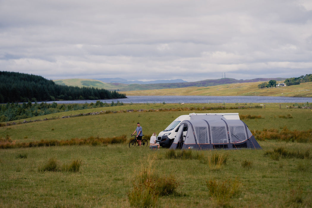 Vango Galli CC Air Inflatable Drive Away Awning - mid lifestyle image of awning side view attached to van with front door half open. grass field in front and hills, tress and lake in background. women sat on coolbox out front and man with bike to the left
