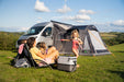 Vango Kela Pro Air Drive Away Awning - Mid lifestyle image of family sat out in front of awning with girl in swimming costume  playing with bubbles. Awning pitched against motorhome with all doors zipped up and curtains in place. background of fields