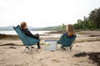 Vango Pinnacle 32L 72Hr Non Electric Camping Cool Box - Grey - lifestyle image, showing two people sat in beach chairs with the cool box on the ground between them, against a beach scene backdrop