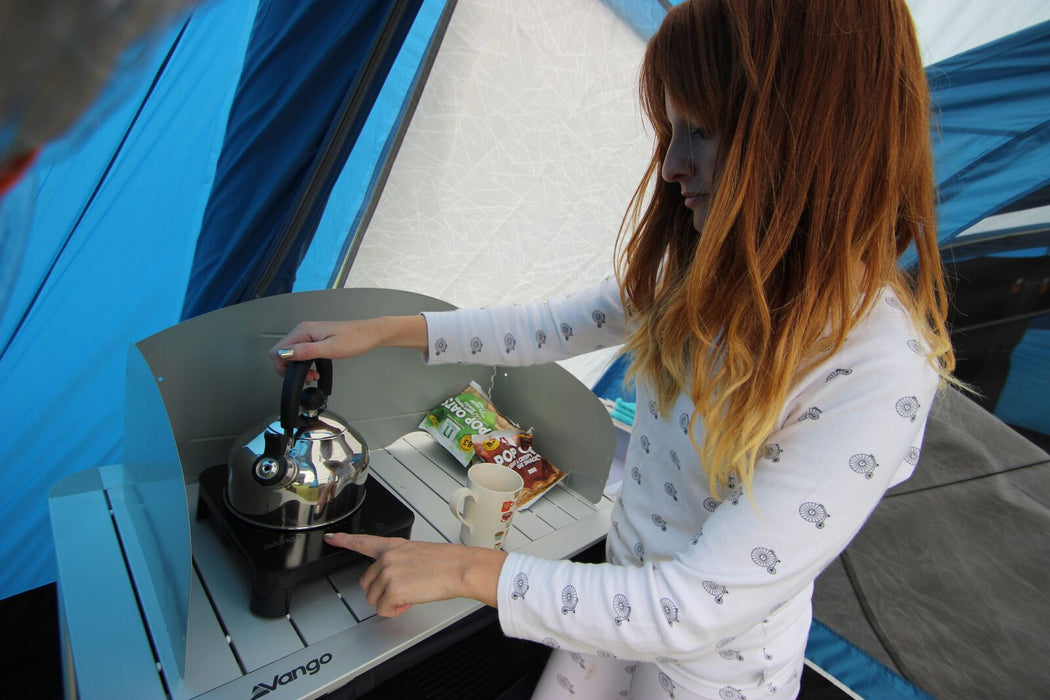 Vango Sizzle Electric Cooker - Black Single 800W Induction Hob lifestyle image of red haired woman with one hand on stainless steel kettle and other on Sizzle hob. In a blue tent with hob on vango kitchen stand with pop oats packets behind mug. 