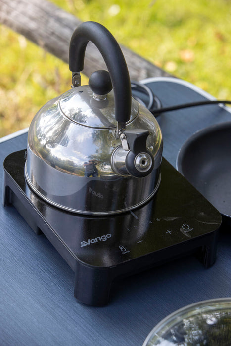 Vango Sizzle Electric Cooker - Black Single 800W Induction Hob lifestyle image of hob on camping table with stainless steel kettle and green grass in background