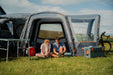 Vango Versos Air Inflatable Cloud Grey Drive Away Awning - Low lifestyle image of the awning pitched and the vehicle on the left with front tunnel door open and couple sat in the main living area with door open. Canopy is pitched on the right with two bikes in the canopy. A table is out in front of the awning to the left and a cool box sits in front of the vango logo on the canopy. The surroundings are green grass and windmills in the background on a hill 