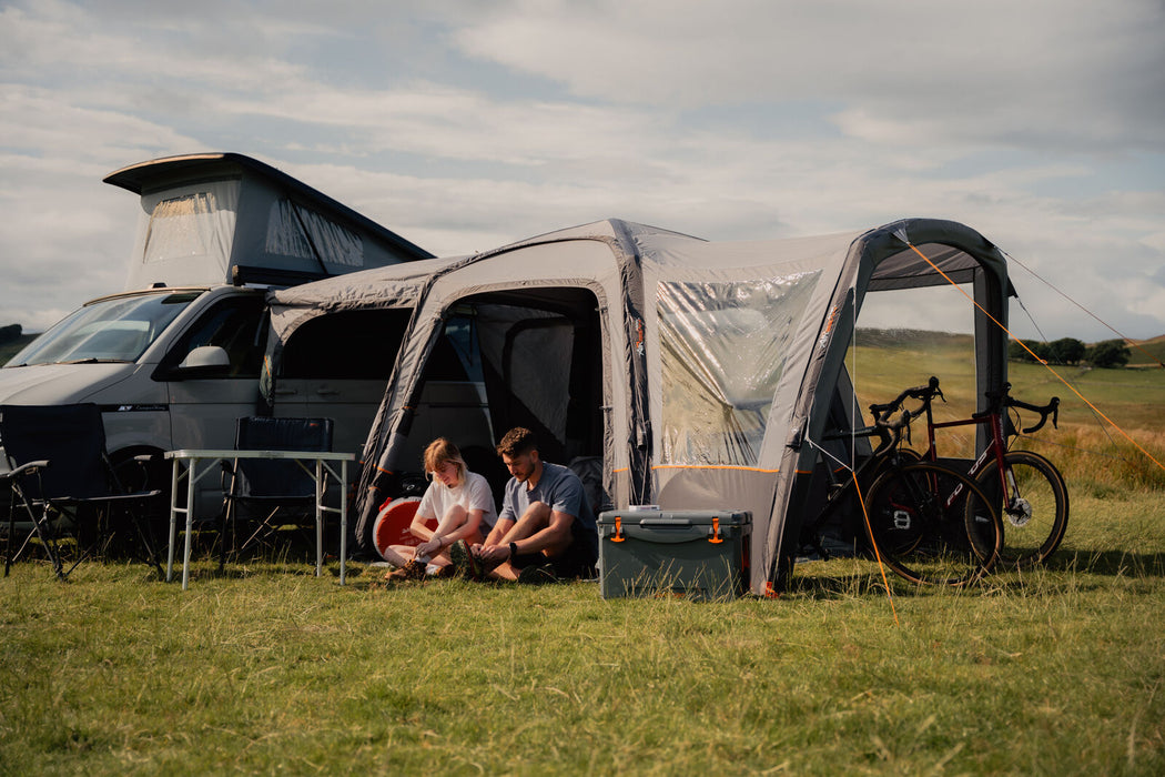 Vango Versos Air Inflatable Cloud Grey Drive Away Awning - Low lifestyle image of the awning pitched and the vehicle on the left with front tunnel door open and couple sat in the main living area with door open. Canopy is pitched on the right with two bikes in the canopy. A table  and chairs are out in front of the awning to the left and a cool box sits in front of the vango logo on the canopy. The surroundings are green grass and a hill with some tress in the background.