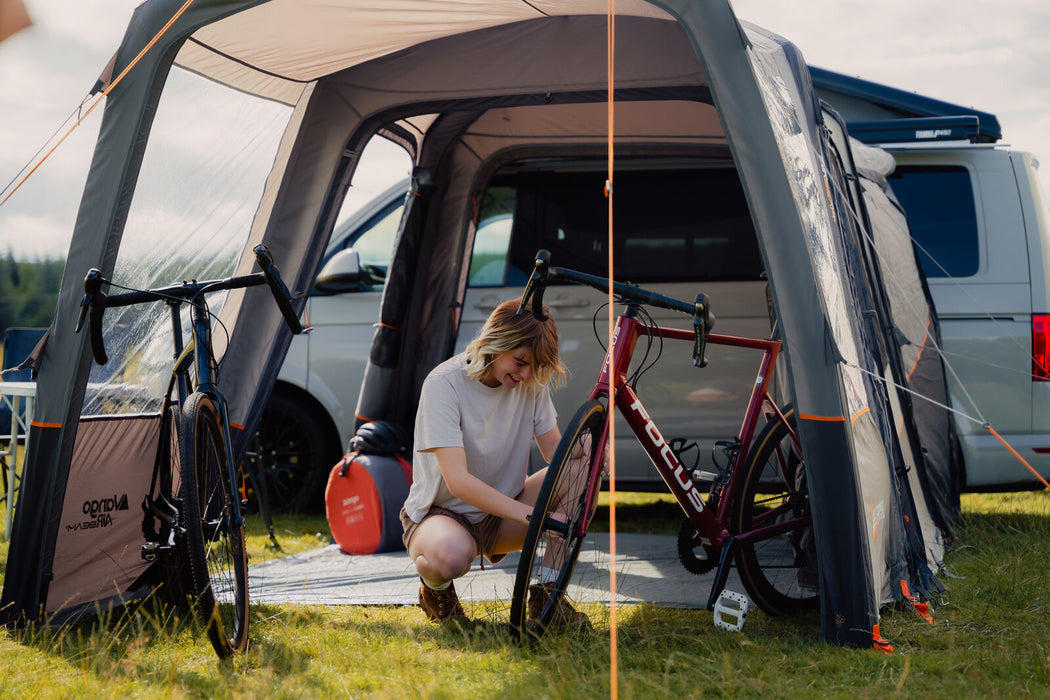 Vango Versos Air Inflatable Cloud Grey Drive Away Awning - Low lifestyle image showing view down the  canopy section to the van. A woman crouches down next to a red bike on the right side in the canop and a black bike is on the left side. A sleeping bag and helmet are in the background in the main living area of the awning.  